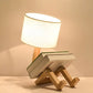 Robot Table Lamp with wooden robot-shaped base holding books and a white fabric drum lampshade providing warm light.
