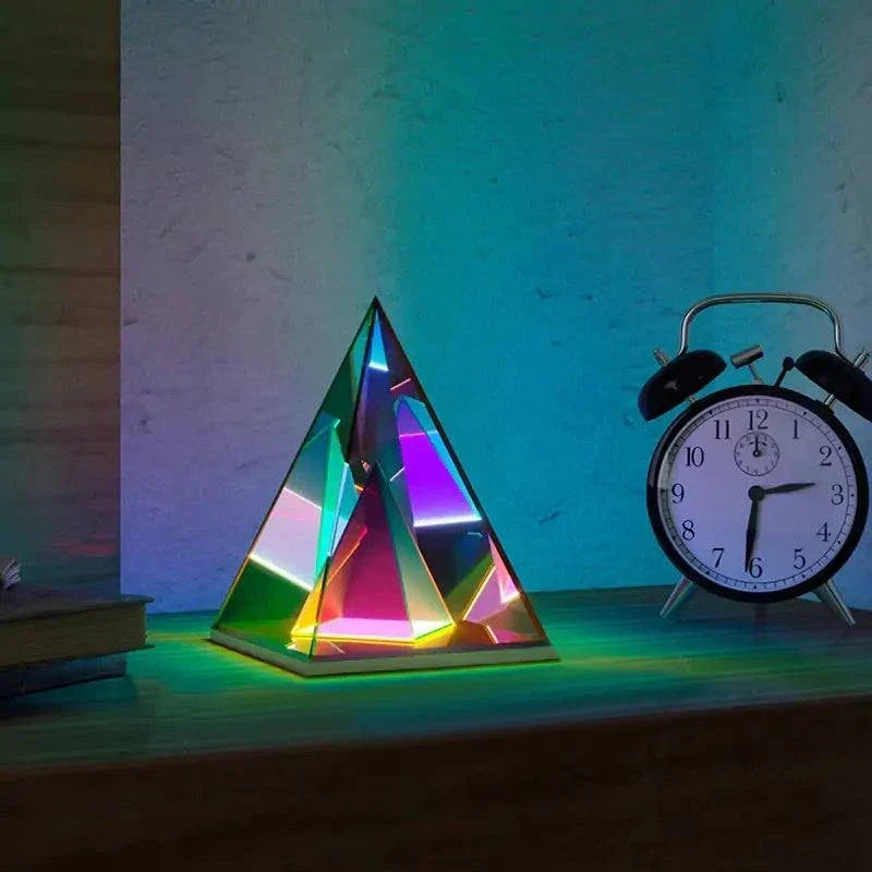 Aura Pyramid Lamp with multicolored, iridescent acrylic pyramid shape emitting ambient glow on wooden surface beside black and white alarm clock