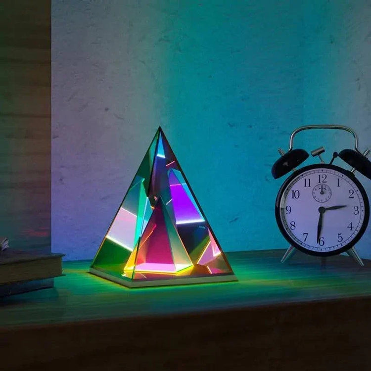 Aura Pyramid Lamp with multicolored, iridescent acrylic pyramid shape emitting ambient glow on wooden surface beside black and white alarm clock
