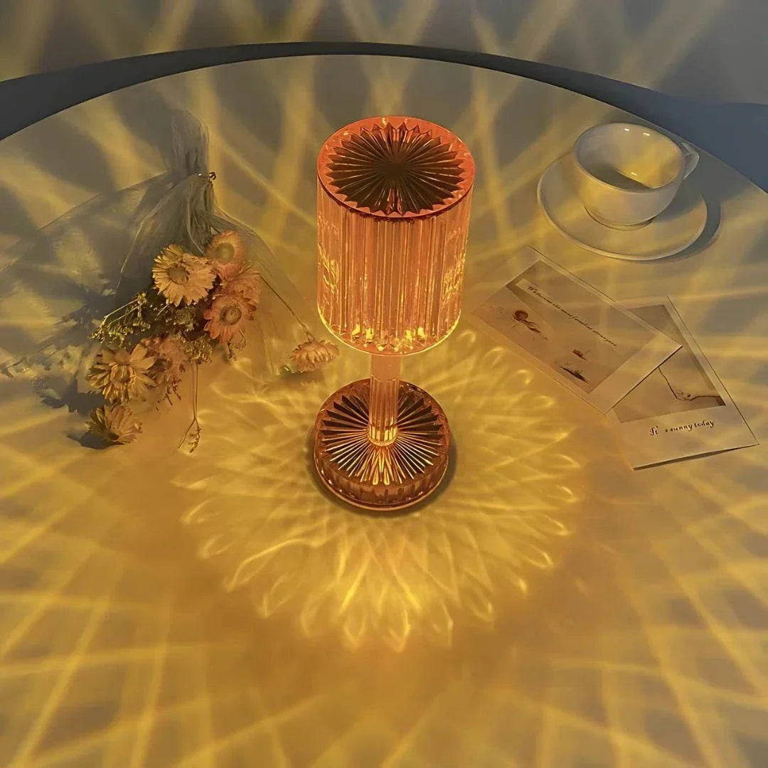 Cristal Lamp with a faceted amber glass body creating intricate light patterns on a white surface, beside dried flowers and a cup