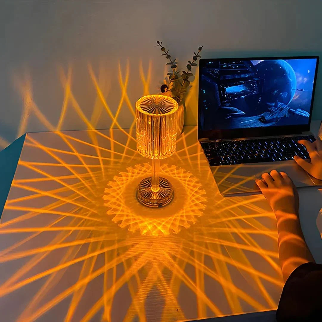 Cristal Lamp with clear faceted glass and a warm amber glow casting intricate radial patterns on a white surface beside a laptop