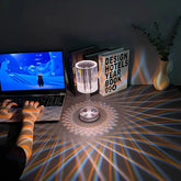 Cristal Lamp with clear faceted glass shade casting intricate light patterns on a wooden table beside books and a laptop