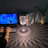 Cristal Lamp with clear faceted glass shade casting intricate light patterns on a wooden table beside books and a laptop
