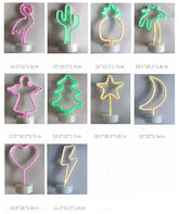 LED Neon Light collection featuring vibrant pink, green, and yellow PVC designs including flamingo, cactus, pineapple, palm tree, angel, Christmas tree, star, moon, heart, and lightning shapes on clear bases