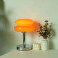 Macaron LED Lamp with a vibrant orange double-oval shade, chrome metal stem and round base, emitting warm light