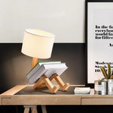 Robot Table Lamp with white fabric shade and wooden base designed to hold books, combining modern and playful style.
