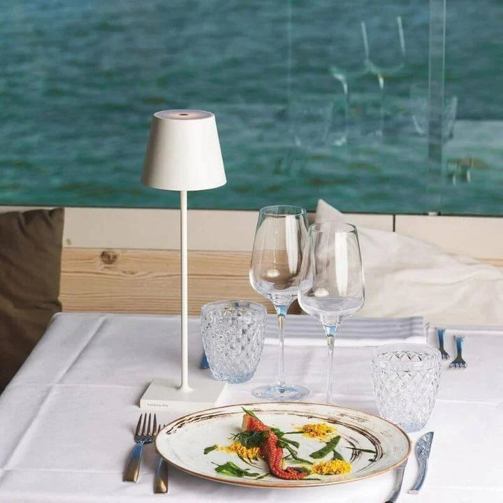 Cordless lamp with a sleek white metal design and minimalist shade, placed on a white tablecloth beside glassware and a decorative plate.