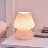 Mushroom LED Lamp in pink hand-blown glass with white diagonal stripes emitting warm light on a white table beside books