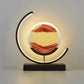 Sandscape Lamp featuring a circular glass vessel with flowing red and beige sand patterns, LED lighting, and a metal arc stand with black base